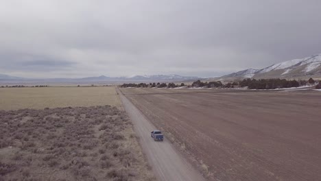 Following-a-truck-as-it-drives-down-a-dirt-road-in-farmland-with-hills-and-mountains-in-the-distance---aerial-view