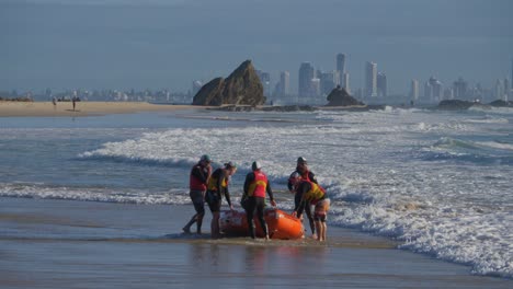 Volunteer-Lifeguards-Pulling-The-Rubber-Boat-On-The-Beach-Shore---Gold-Coast-Skyline-From-Currumbin---Gold-Coast,-Queensland,-Australia