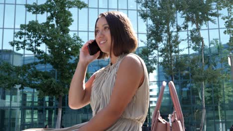 Elegant-attractive-businesswoman-is-making-a-phone-call-in-bright-sunshine-in-front-of-the-tall-glass-front-of-a-modern-office-building-with-isolated-green-trees