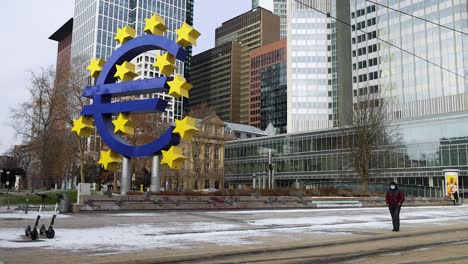 Hombre-Adulto-Con-Mascarilla-Durante-La-Pandemia-Frente-A-La-Escultura-Del-Signo-Del-Euro-En-Willy-brandt-platz-En-Invierno-En-Frankfurt-Am-Main,-Alemania