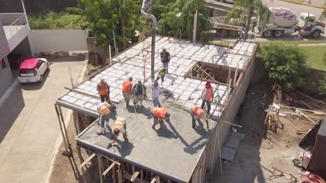 Vista-Aérea-De-Los-Trabajadores-De-La-Construcción-Que-Trabajan-En-El-Techo-Vertiendo-Cemento-Para-Construir-Una-Estructura-De-Techo-O-Piso-Usando-Chalecos-Naranjas