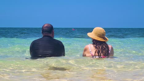 Couple-Sitting-Together-In-Ocean-As-Waves-Gently-Go-Past-Them-Near-Beach