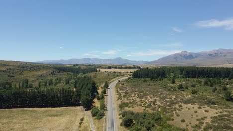 Aerial-lowering-on-a-lonely-road-surrounded-by-pine-tree-woods-with-mountains-in-background