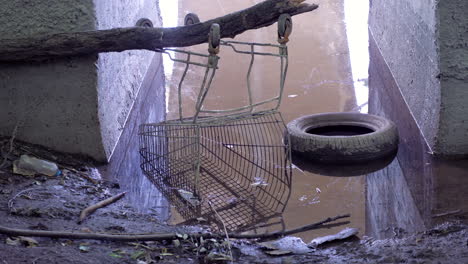 Rusted-Discarded-Shopping-Trolley,-Car-Tire-At-River-Bank