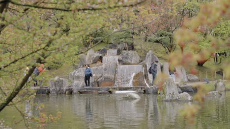 Peering-through-branches-at-the-reflection-of-a-cascading-waterfall-in-a-public-Japanese-garden,-slow-motion