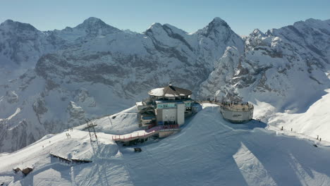 Aerial-of-ski-lift-building-with-busy-terrace-on-top-of-snow-covered-mountain
