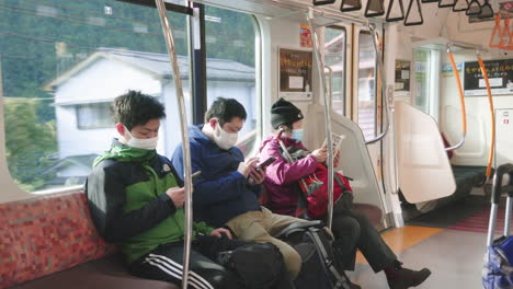 Viajeros-Japoneses-Con-Máscaras-Usando-Teléfonos-Inteligentes-Y-Leyendo-Libros-En-El-Tren-En-Okutama,-Japón