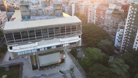 Aerial-rising-over-brutalist-style-National-Library-surrounded-by-trees-and-buildings-at-golden-hour,-Buenos-Aires