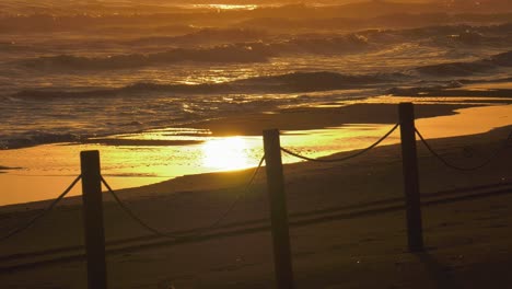 Sandy-beach-with-waves-breaking-onto-shore-reflecting-sunrise,-mediterranean-sea,-coastal-view,-spain