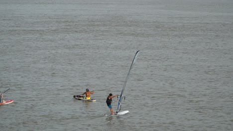 Hombre-Haciendo-Windsurf-En-El-Río-Han-Pasando-Por-Otros-Sentados-En-Tablas-De-Surf-Y-Remando-En-El-Agua