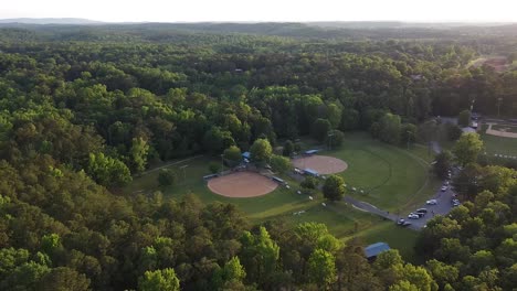 Baseball-and-softball-field-in-the-woods-surrounded-by-mountains-at-sunset,-aerial