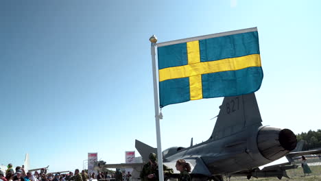 Bandera-Sueca-Ondeando-Junto-A-Saab-Jas-39-Gripen-De-La-Fuerza-Aérea-Sueca-En-El-Aeropuerto-De-Gdynia,-Polonia-Con-Pilotos-De-Combate-Y-Espectadores-En-Tierra