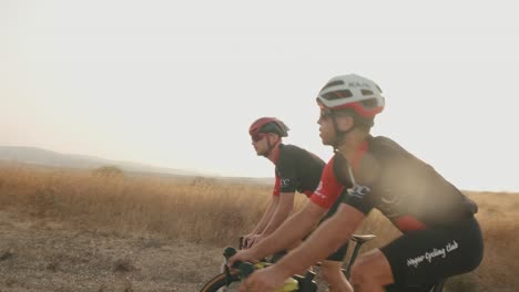Dos-Profesionales-Ciclistas-De-Carretera-En-Cámara-Lenta-Montando-En-La-Carretera-Vacía-Del-Desierto-Mientras-El-Sol-Enciende-La-Cámara