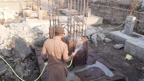 Ein-Mann-Im-Pathani-Anzug-Und-Mit-Schutzbrille-Gibt-Wasser-Auf-Den-Untergrund-Der-Baustelle,-Um-Ihn-Zu-Stärken-–-Bewässerung-Auf-Einer-Neuen-Baustelle-In-Indien