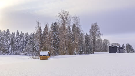 Zeitraffer-Einer-Wunderschönen-Winterlandschaft-Mit-Fliegenden-Wolken-Und-Sonnenlicht-Am-Himmel---Fasssauna-Und-Holzhaus-Auf-Einem-Verschneiten-Feld-Vor-Weißen-Tannen