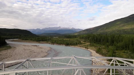 Drone-flying-over-a-bridge-above-the-turquoise-waters-of-Toad-River-in-the-natural-landscape-of-northern-British-Columbia,-Canada