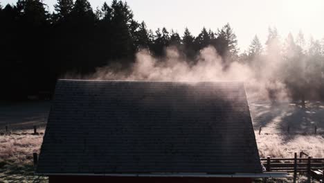 Mist-rising-from-the-roof-of-a-red-barn-as-the-morning-sun-warms-it,-aerial-orbit