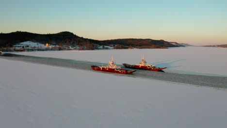 Aerial-view-of-two-car-ferries-passing-as-they-cross-a-snow-covered-river-at-sunset