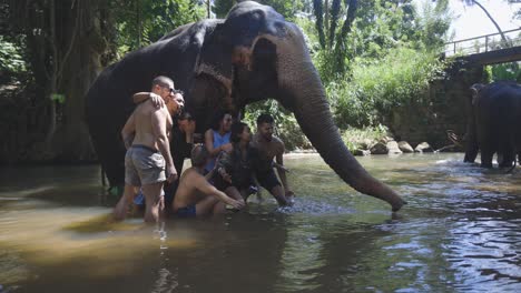 Statische-Aufnahme-Glücklicher-Touristen,-Die-Auf-Einen-Elefanten-Warten,-Um-Sie-Mit-Wasser-Zu-überschütten,-Und-Im-Hintergrund-Ein-Elefant,-Der-Im-Fluss-Läuft,-Mirissa,-Sri-Lanka