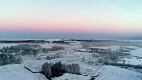 Sunset-over-a-rural-landscape-of-forests,-fields-and-roads-covered-with-snow-and-mist-in-winter
