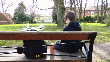 Boy-With-Drone-Eating-Lunch-on-a-Bench-in-a-Park,-Pedestrians-Walking-By