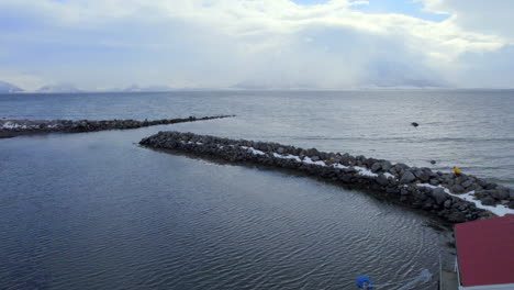 Low-aerial-dolly-shot-of-the-Ase-harbor-and-breakwater-going-out-over-the-ocean-with-distant-snow-covered-mountains,-Norway