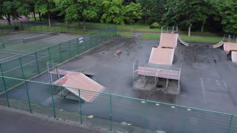 Aerial-view-flying-above-fenced-skateboard-park-ramp-and-tennis-court-in-empty-closed-public-playground