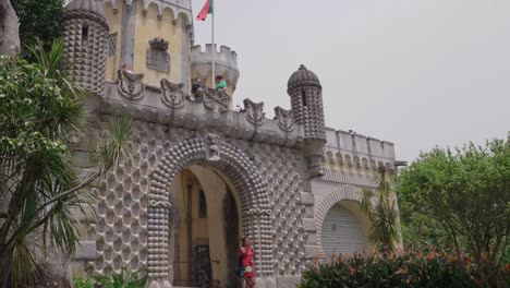 Dame-In-Rot-Verlässt-Die-Architektonische-Hauptfassade-Des-Schlosses-Von-Sintra-Pena,-Mit-Unglaublich-Detailliertem-Beton-Und-Gelber-Struktur-Mit-Wehender-Portugiesischer-Flagge-Oben,-Sintra,-Portugal,-Lissabon