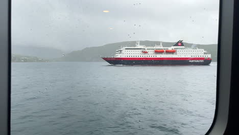 Static-shot-through-a-ferry-window-passing-worlds-famous-Hurtigruten-in-Norway-on-a-rainy-moody-day