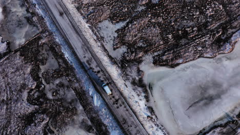Aerial-Top-Down-View-Tracking-Van-Car-Driving-Moving-in-Winter-on-Road-Through-Wild-Snowy-Rocky-Lands-and-Frozen-Wetlands-in-Búlandstindur-Mountain-Area,-Iceland