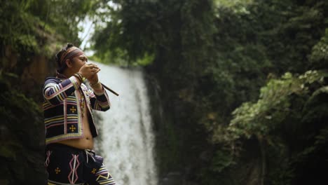 Indigenous-person-playing-Kubing-tribal-instrument,-behind-is-a-waterfall
