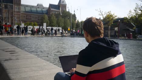 Un-Joven-Apuesto-Sentado-A-Orillas-Del-Canal-Frente-Al-Rijksmuseum-Con-El-Cartel-I-Amsterdam-Escribiendo-Un-Texto-En-Una-Laptop