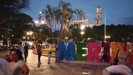 Empujando-El-Cartel-De-Merida-Para-Que-La-Gente-Se-Tome-Selfies-Frente-A-La-Catedral-De-San-Ildefonso-Al-Fondo,-Al-Atardecer