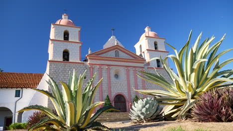 Die-Historische-Fassade-Des-Spanischen-Katholischen-Missionsgebäudes-Santa-Barbara-In-Kalifornien-Zwischen-Saftigen-Einheimischen-Pflanzen-Gleitet-Nach-Rechts