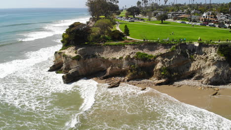 Aerial-drone-shot-of-green-beach-cliffs-over-the-ocean-waves-crashing-on-the-sandy-coast-in-Santa-Barbara,-California
