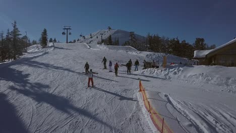 Luftaufnahme-Einer-Gruppe-Von-Skifahrern-Neben-Einem-Holzhaus-Auf-Einer-Skipiste-Und-Einer-Von-Ihnen-Beginnt-Voller-Freude-Mit-Dem-Skifahren-Und-Winkt-An-Einem-Sonnigen-Tag-In-Die-Kamera,-Mit-Wunderschönen-Bergen-Im-Hintergrund