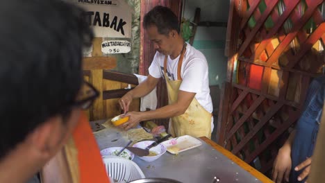 A-street-vendor-wearing-a-bright-yellow-apron-begins-making-a-fresh-order-of-martabak,-a-stuffed-pancake-and-popular-street-food-in-Asia-and-the-Middle-East