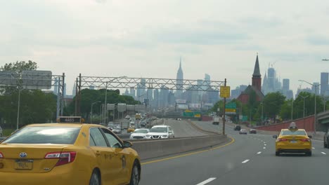 Skyline-of-the-skyscrapers-of-Manhattan-with-yellow-cabs-in-the-foreground,-filmed-from-an-approaching-car-in-slow-motion-on-495-Highway-in-Queens,-New-York-City