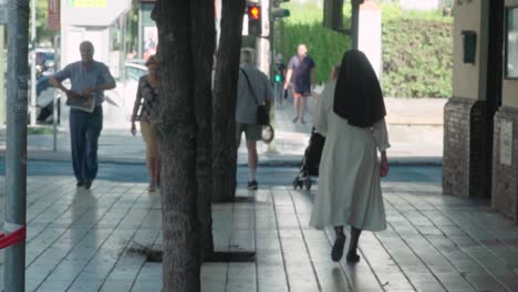Nun-walking-by-people-on-sidewalk-in-Seville,-Spain,-view-from-behind