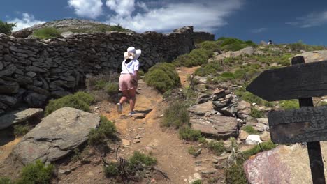 Girl-in-summery-hiking-outfit-and-hat-walking-up-path-next-to-wall-on-mountain,-camera-right-pans-to-direction-signs