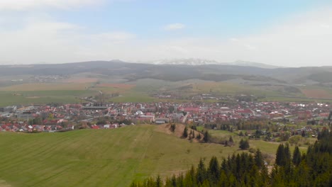 Forward-aerial-of-small-town-during-the-day-with-forest-in-foreground-and-mountains-in-distance