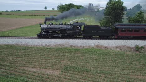 Vista-Aérea-De-Un-Motor-De-Vapor-Antiguo-Con-Automóviles-De-Pasajeros-Resoplando-A-Lo-Largo-De-La-Campiña-Amish