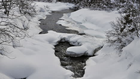 Time-lapse-of-a-small-stream-in-the-French-Alps-that-has-snow-covered-sides-and-bank