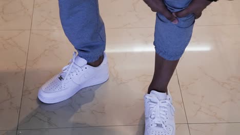 Close-up-of-Male-legs-rolling-up-the-hem-of-his-blue-pants-wearing-white-sneakers-standing-up-on-a-beige-floor---interior-scene-tripod-shot