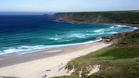 Ocean-waves-along-the-wide-sandy-shore-at-Sennen-Cove-in-Cornwall-with-surfers-and-tourists-walking-on-the-beach