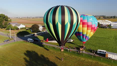 A-large-hot-air-balloon-lands-and-maneuvers-into-position-in-a-green-meadow-with-surrounding-Amish-farmland,-aerial-drone-view