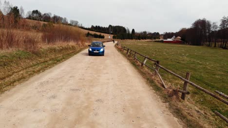 A-tracking-drone-shot-following-a-blue-car-driving-in-the-countryside-beside-some-fields