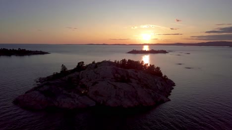 Rocky-Island-with-Pine-Trees-in-Blue-Lake-at-Sunset,-Drone-Aerial-Wide-Orbit-Pan-Pedestal-Down