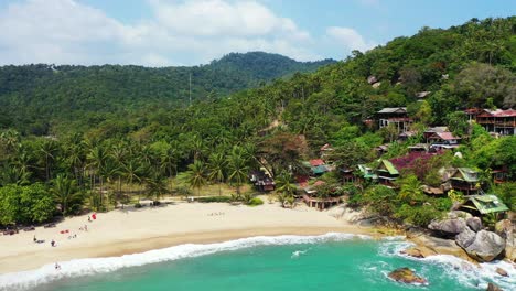Elevated-waterfront-bungalows-in-the-palm-forest,-foamy-waves-washing-sandy-beach-on-tropical-Thailand-island