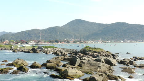 Aerial-cinematic-view-rocks-coastline-and-boats,-Praia-Armacao,-Florianopolis,-Santa-Catarina,-Brazil